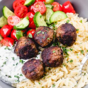 Greek meatballs with orzo, tzatziki sauce, and cherry cucumber salad in a grey bowl with a fork.