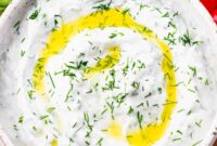 Tzatziki sauce, topped with olive oil and chopped dill, in a white bowl and vegetables and pita bread around it.