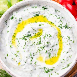 Tzatziki sauce, topped with olive oil and chopped dill, in a white bowl and vegetables and pita bread around it.